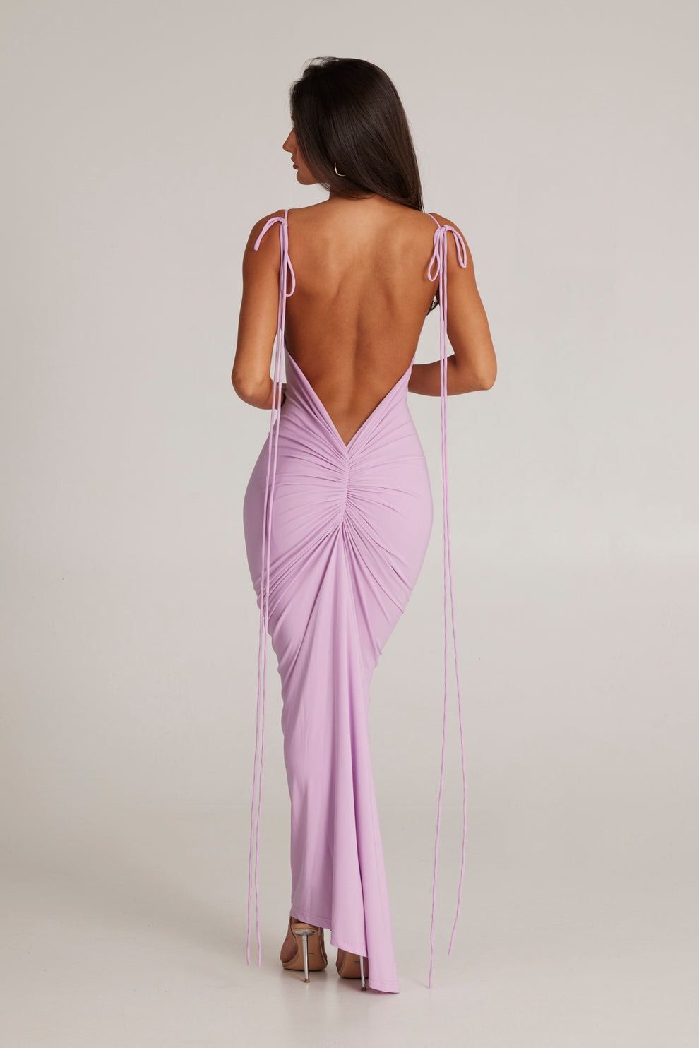 Woman in a lilac plunging backless maxi dress with delicate spaghetti straps and ruched detailing.